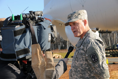 A man in a military uniform stands outdoors and talks into a microphone that is being held by a person who is obscured by a large video camera.  In the background is a large white blimp.