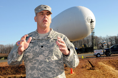 A man in a military uniform is standing outdoors with his hands outstretched.  Behind him is a a large, white blimp, which is still on the ground and attached to a mast. 