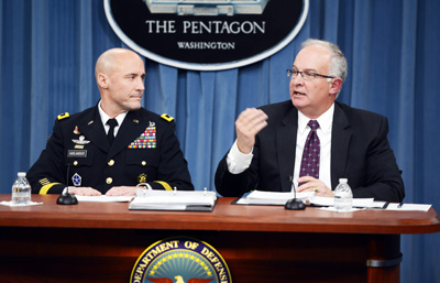 A man in a military uniform and a man in a suit sit together at a wooden table.  The table has a seal on the front that says "Department of Defense."  On the wall behind them are blue drapes and a sign that says "The Pentagon."
