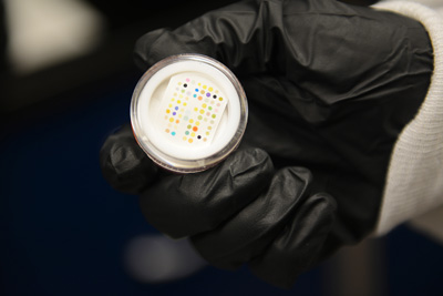 A gloved hand holds a small plastic disc that contains a piece of paper with an array of colored dots.
