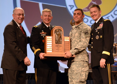 Four men, one in a suit and three in military uniforms stand next to each other on a stage.  Two of the men hold a large wooden trophy.