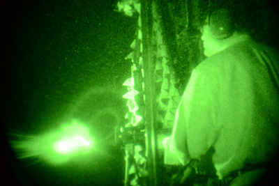 A man fires a rifle.  The entire scene is green, as it appears though night vision goggles.