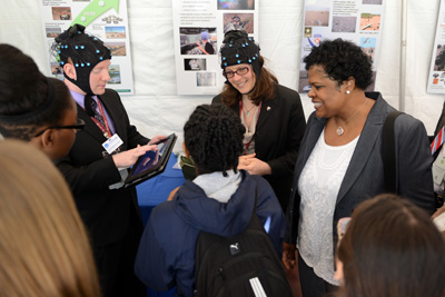 A woman and a man wear stocking caps that appear to have integrated sensors.  They speak with children. A woman who appears to be a teacher stands nearby.
