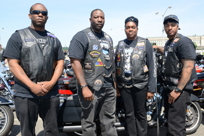 Four men in leather vests stand in front of their motorcycles.