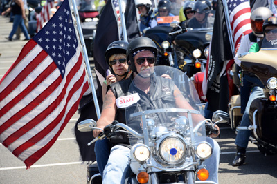 A man and a woman ride a motorcycle.  An American flag is attached to the bike.  Nearby are other bikers.