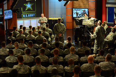 Dozens of uniformed individuals are seated in a large room.  At the front of the room a man in military uniform stands and faces them.  Behind the man is a large U.S. Army logo and several television screens. At the front of the room is a uniformed man operating a large television camera.