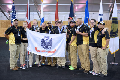 A dozen individuals in black and yellow shirts stand together and hold a U.S. Army flag.  Behind them are an array of other flags.