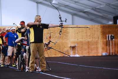 A man in a yellow and black shirt draws back the string on a bow and aims an arrow.