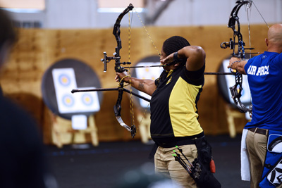 A man in a blue shirt, and a woman in a black and yellow shirt, drawing back a bow to shoot an arrow at a down-range target.