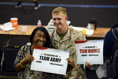 A woman holds a sign that says "Team Army."  Next to her a young man in a military uniform holds a sign that says "Team Marines."