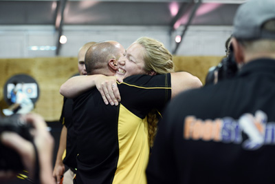 A man and a woman hug each other in celebration.  Others stand nearby and watch. Some have cameras.