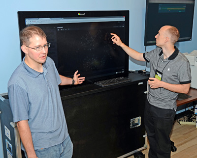 Two young men stand in front of a television, which display graphical information about computer networks.