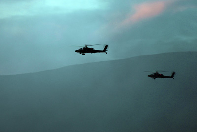 Two helicopters fly near each other in front of a mountain range.  The sky is hazy and tinged blue and red from the sunset.