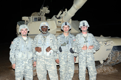 At night time, four men in military uniforms stand in front of a tank.