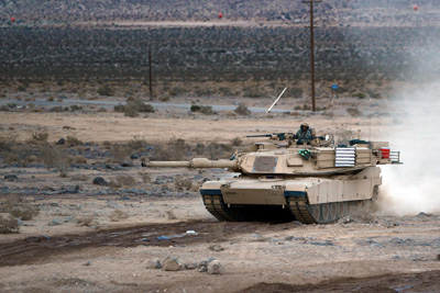 A military tank kicks up dust as it rolls across a barren landscape. Its turret is swiveled to its right.