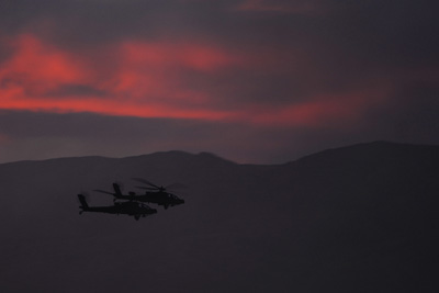 Two helicopters fly near each other in front of a mountain range.  The sky is hazy and tinged red from the sunset.