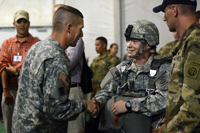 A young male Soldier wearing a helmet shakes hands with an older male Soldier. Soldiers and civilians stand nearby.