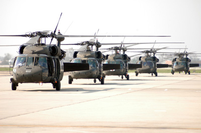 Five military helicopters are lined up on a large concrete surface.