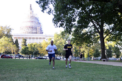 Two individuals run along a grassy field. In the background is the U.S. Capitol building.