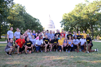 Around two dozen individuals wearing fitness clothing pose together on a grassy field. In the background is the U.S. Capitol building.