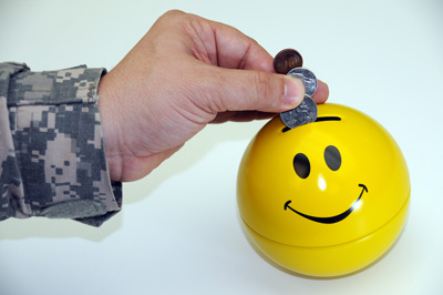 The hand of a person in a military uniform inserts coins into a ball-shaped yellow bank that is painted with a smiley face.