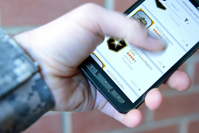 The hand of a uniformed individual holds a cell phone.  The screen displays the names of cell phone applications.  A brick wall looms in the background.