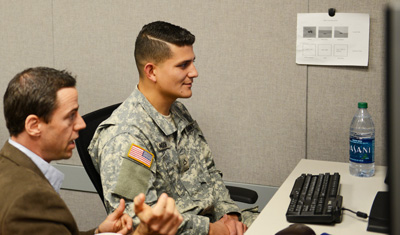 A young man in a military uniform sits at a computer. A civilian man sits next to him.  