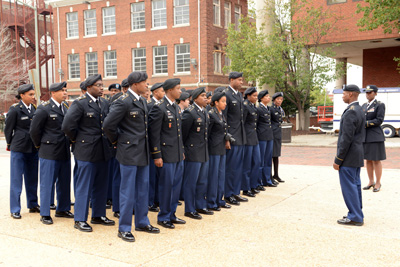 Dozens of young people in military uniforms stand in formation, outdoors, near college campus buildings.  Another uniformed person stands in front of them.