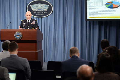 A man in a military uniform stands behind a lectern.  Hanging from blue drapes behind him is an oval-shaped sign that says "The Pentagon," and which features a line drawing of the Pentagon.  To his left is a television that displays a pie chart. People are seated in front of him.