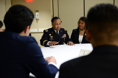 A man in a military uniform sits at a table next to a woman. Other people are seated at the table as well. He is talking to them.