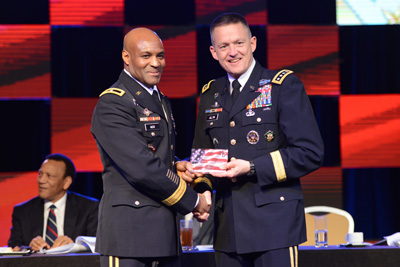 Two men in military uniforms stand alongside each other.  They are holding what appears to be a plaque with the image of an American flag on it. 