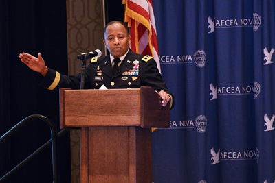 A man in a military uniform stands behind a lectern.  His hand is outstretched.  Behind him is an American flag and drapery with the words "AFCEA NOVA" written on them.