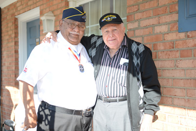 Two men, both wearing hats that indicate their status as veterans, stand near each other. One has his arm on the shoulder of the other.
