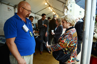 A man in a blue shirt with an artificial arm shakes hands with a woman in colorful blouse.  The two are standing under a tent.