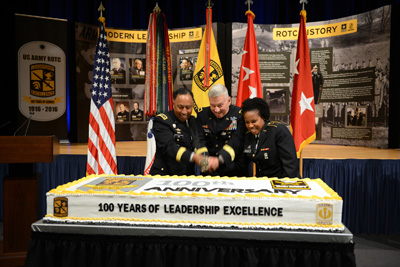 Two men and one woman in military uniforms use a sword to cut a large cake.  The cake reads "100 years of leadership excellence."  In the rear is an array of flags and display boards which relay information related to ROTC.