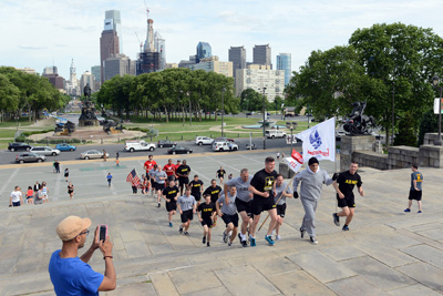 Dozens of individuals in athletic clothing run up a set of stone stairs.  In the background looms the skyline of Philadelphia.