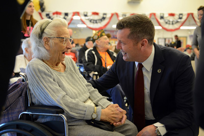 A man in a suit kneels down next to an elderly woman in a wheelchair and talks with her.