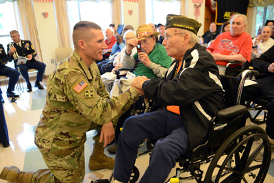 A man in a military uniform, down on one knee, speaks to an elderly man in a wheelchair.  Nearby, others are seated.
