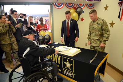 An elderly man in a wheelchair uses a sword to cut a cake, which sits on a table bearing the words "U.S. Army."  Behind the table are a man in a suit, and another in a military uniform.