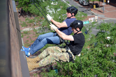 Two men, one in a military uniform, rappel down the side of a building.