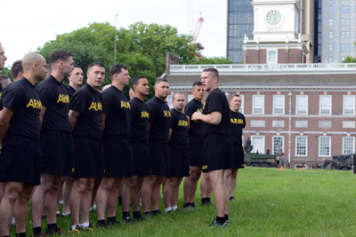 More than a dozen men in black T-shirts that say "Army" stand in formation. Another man, similarly dressed, stands in front of them and talks with them.  In the background is a historical building.