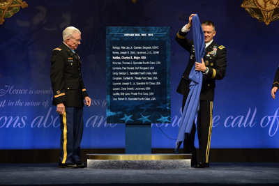 Two men in military uniforms stand on a stage and flank a placard, which bears the names of service members.  One holds a blue sheet.