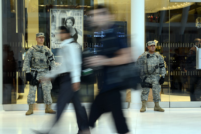 Two Soldiers stand against glass walls. Civilians walk by quickly.