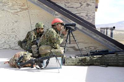 One soldier kneels and looks through a scope, while another sits nearby.