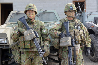 Two military personnel stand near one another. They carry rifles.