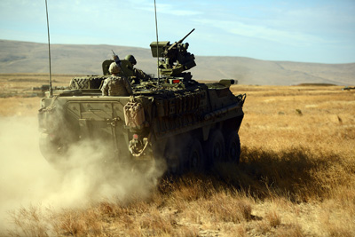 A military combat vehicle kicks up dust in a dry, dusty field environment.  Mountains loom in the background.