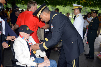 A elderly man is seated in a wheelchair.  He wears a hat with the word "Korea" on it.  A man in a military uniform places a medal around his neck.