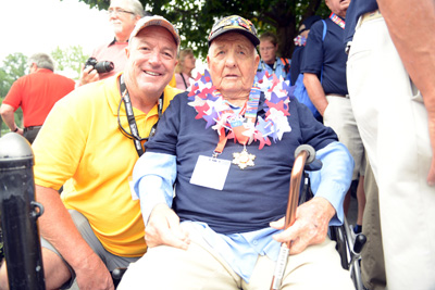 An older man sits in a wheelchair with a cane in one hand and a medal around his neck. Another man kneels beside him.