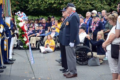 Two elderly men stand at attention, outdoors, in front of a wreath.  Around them military personnel and dozens of civilians observe.