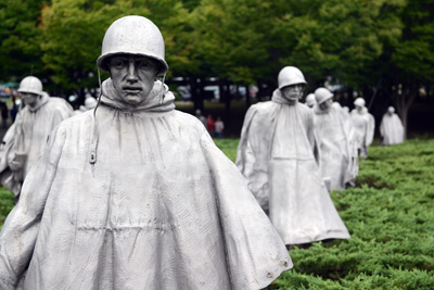 Nearly a dozen statues depicting soldiers who are wearing ponchos and helmets, stand amidst greenery.